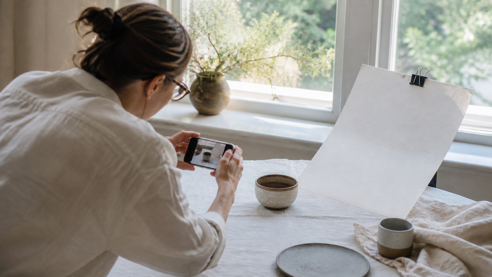 Emprendedora fotografía productos cerca de una ventana con el celular